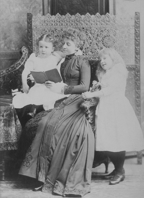 A mother in a long dress reading to two small children on a carved chair, Montreal, 1891